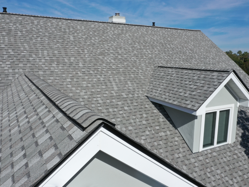 A rooftop with gray asphalt shingles and a small dormer window. The sky is clear and blue, emphasizing the roofs texture and pattern.