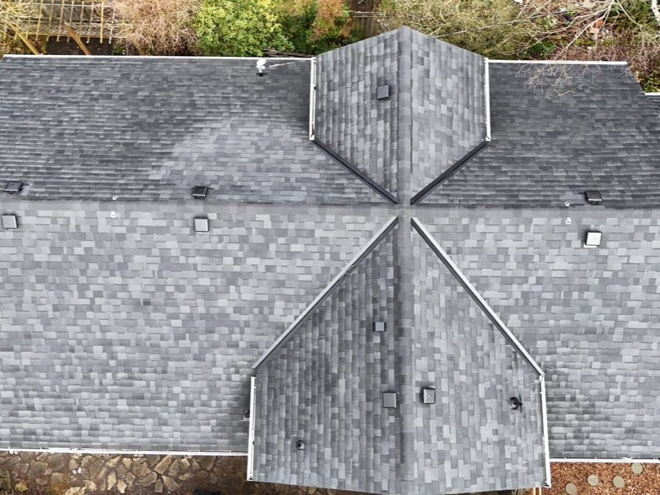 Aerial view of a gray shingled house roof with intersecting gables and several vents, surrounded by trees and some landscaping visible at the edges.