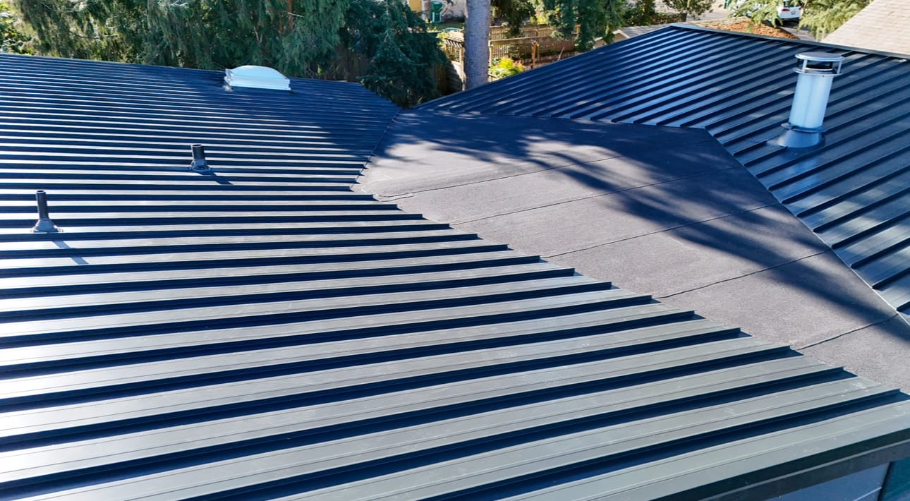 Aerial view of a modern black metal roof with ridges and vent pipes. The design features intersecting sections, and theres a chimney vent on one side. Sunlight casts shadows from nearby trees onto the roofs surface.