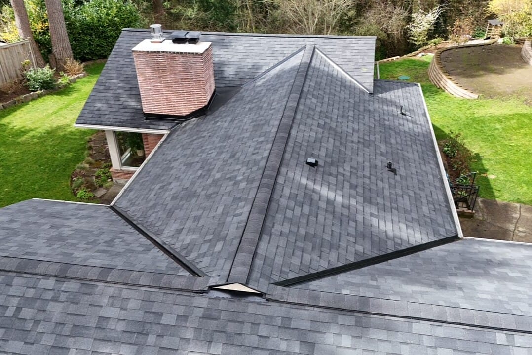 Aerial view of a house with a dark gray shingle roof, brick chimney, and surrounding green lawn with trees, shrubs, and a curved landscaped area in the backyard.