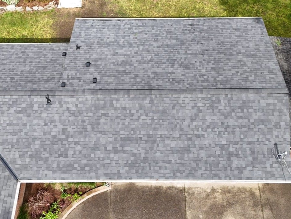 Aerial view of a house roof with gray asphalt shingles in good condition, surrounded by a driveway, grass, and some trees.
