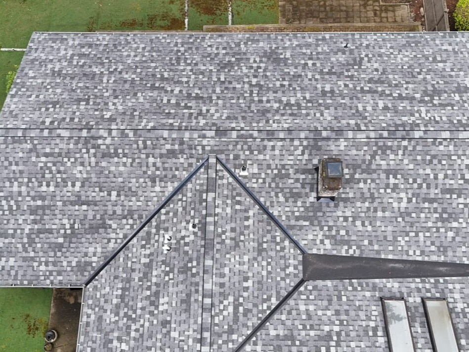 Aerial view of a house roof with gray asphalt shingles, a skylight, and clean, geometric lines. Surrounding green grass and partial view of a paved area are visible.