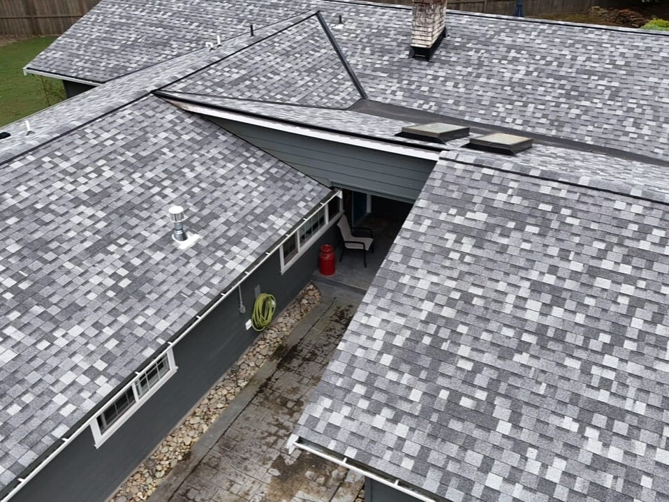 Aerial view of a house with fresh, gray asphalt shingles on the roof, multiple intersecting rooflines, skylights, a chimney, and a small patio area below, surrounded by landscaping.