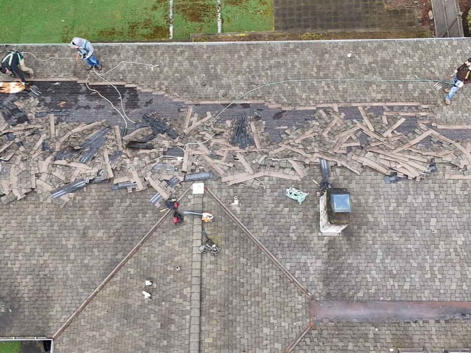 Aerial view of three workers removing old shingles from a roof, revealing damaged wood underneath. Debris is scattered across the roof, and the workers are using ropes and tools for safety and repair.