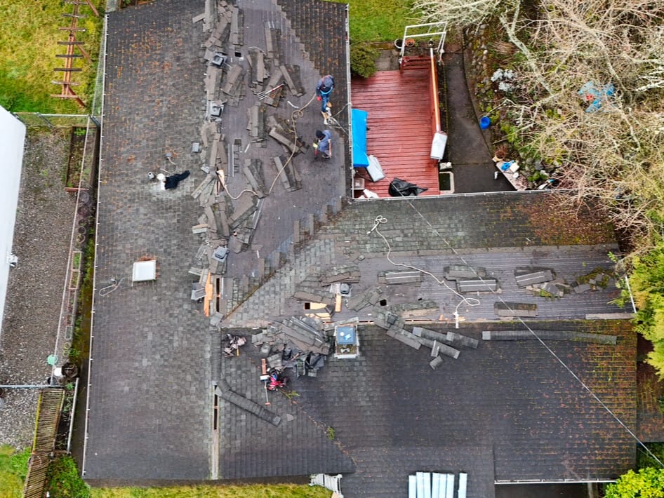 Aerial view of a house roof with workers removing old roofing materials. The roof appears worn and shingles are scattered. Nearby, a backyard deck with materials and tools visible. Trees surround the property.