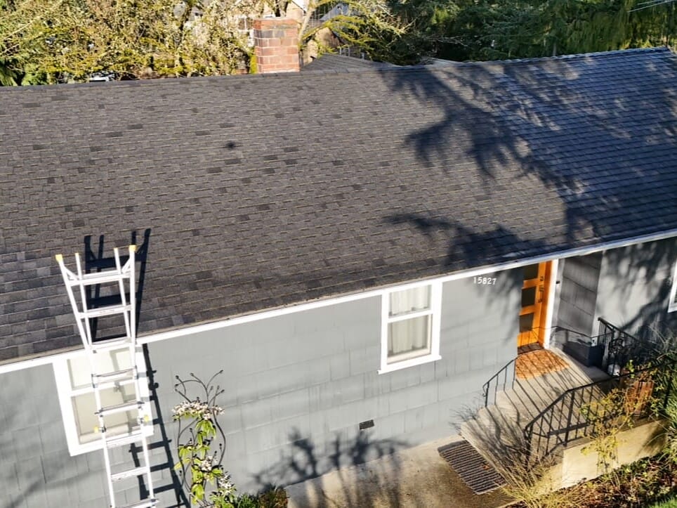 A gray house with a shingled roof, orange front door, and white-framed windows. A tall metal ladder leans against the roof near a chimney. Trees cast shadows on the roof and house.
