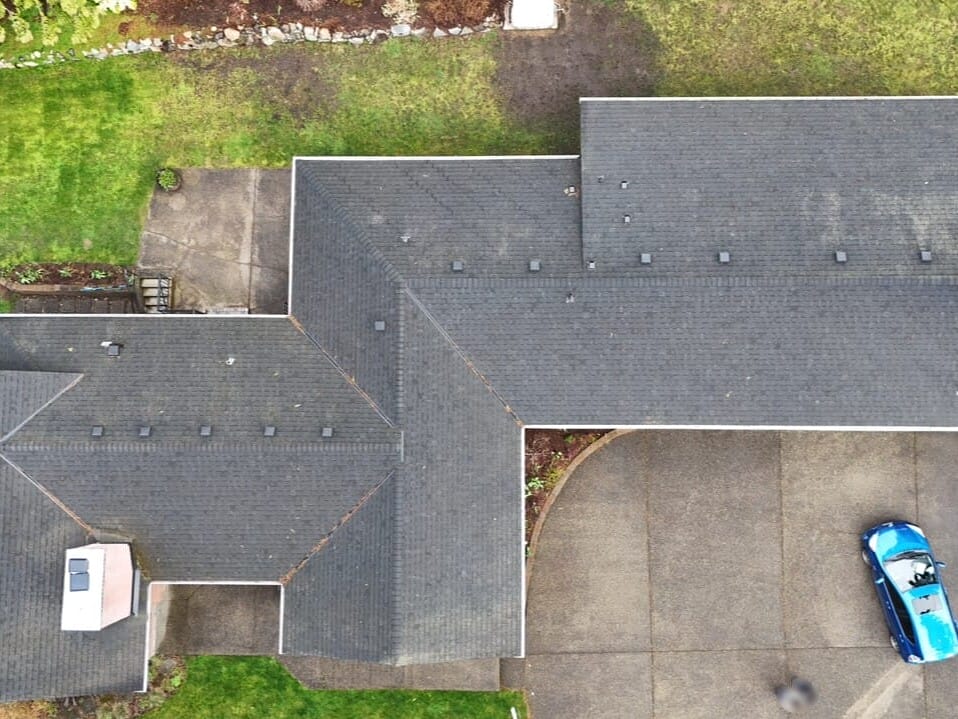 Aerial view of a house with a dark gray roof, surrounded by green lawn and trees. A blue car is parked on the driveway to the right of the house. The yard is bordered by shrubs and a stone-lined garden.