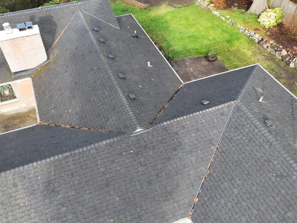 Aerial view of a house with dark gray shingle roofing, surrounded by green grass, landscaping, and a wooden fence along the backyard.