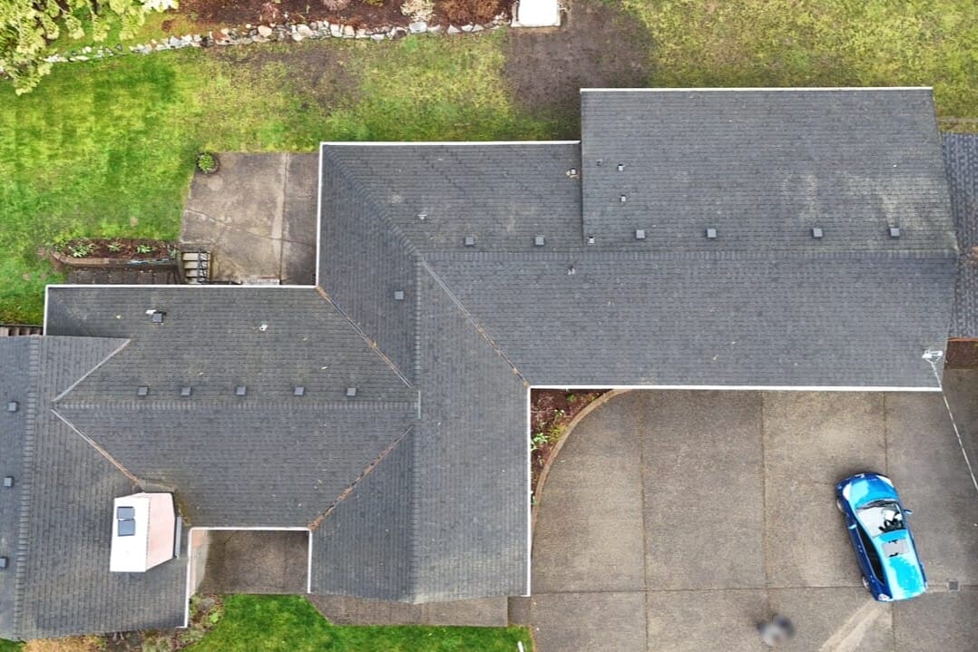 Aerial view of a house with a dark gray roof, surrounded by green grass and trees. A blue car is parked on the driveway next to the house. The scene includes paved walkways and landscaped areas.
