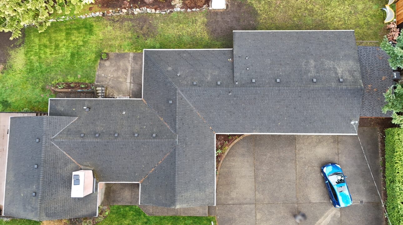 Aerial view of a house with a dark gray roof, surrounded by green grass and trees. A blue car is parked on the driveway next to the house. The scene includes paved walkways and landscaped areas.