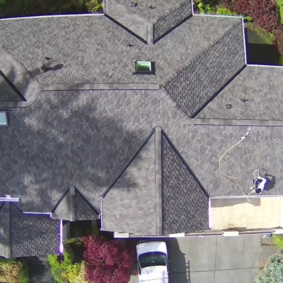 Aerial view of a house with an asphalt shingle roof featuring multiple gables and a skylight. A person is working near the edge. A parked white vehicle is visible in the driveway surrounded by colorful trees and greenery.