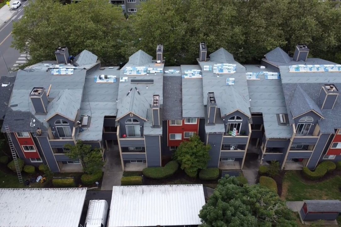 Aerial view of a multi-story apartment building with a gray roof in progress of being reroofed. The building has blue and red siding, surrounded by trees. Several stacks of roofing materials are visible on the rooftop.