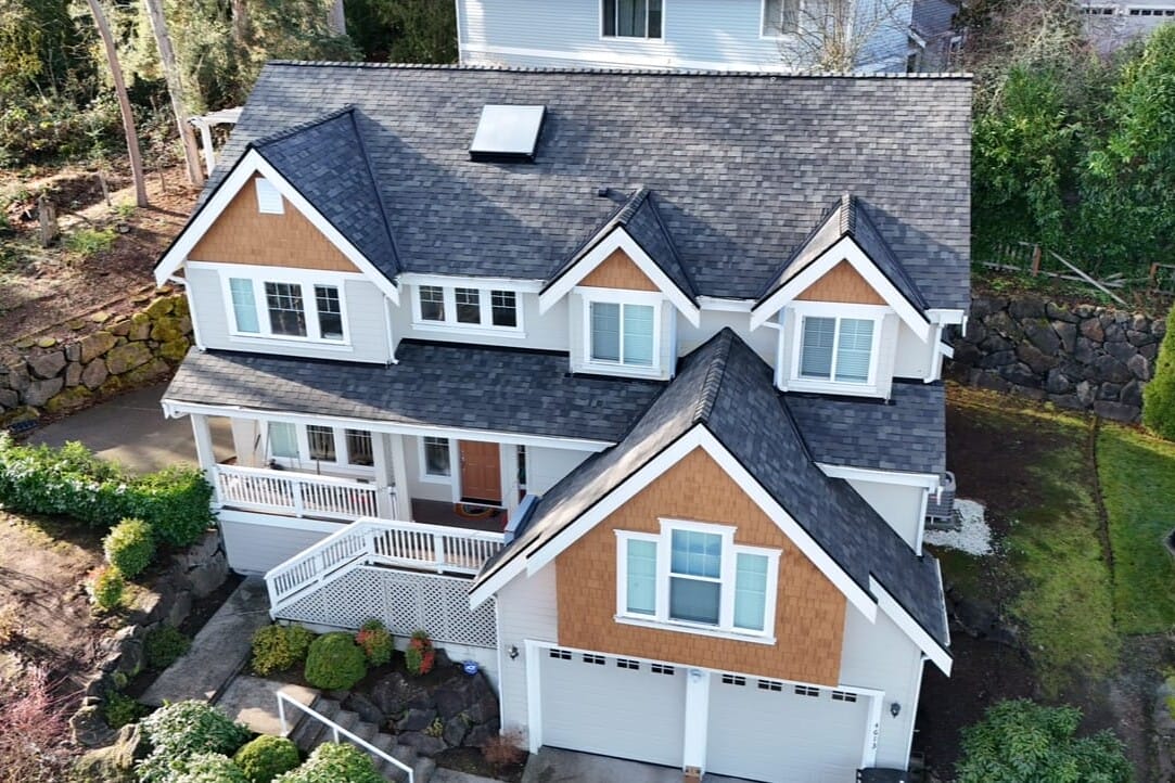 Aerial view of a two-story house with a gray roof, white siding, and brown accents. The house has multiple gables, a skylight, and a front porch. The garden includes shrubs and stone paths, and theres a two-car garage.