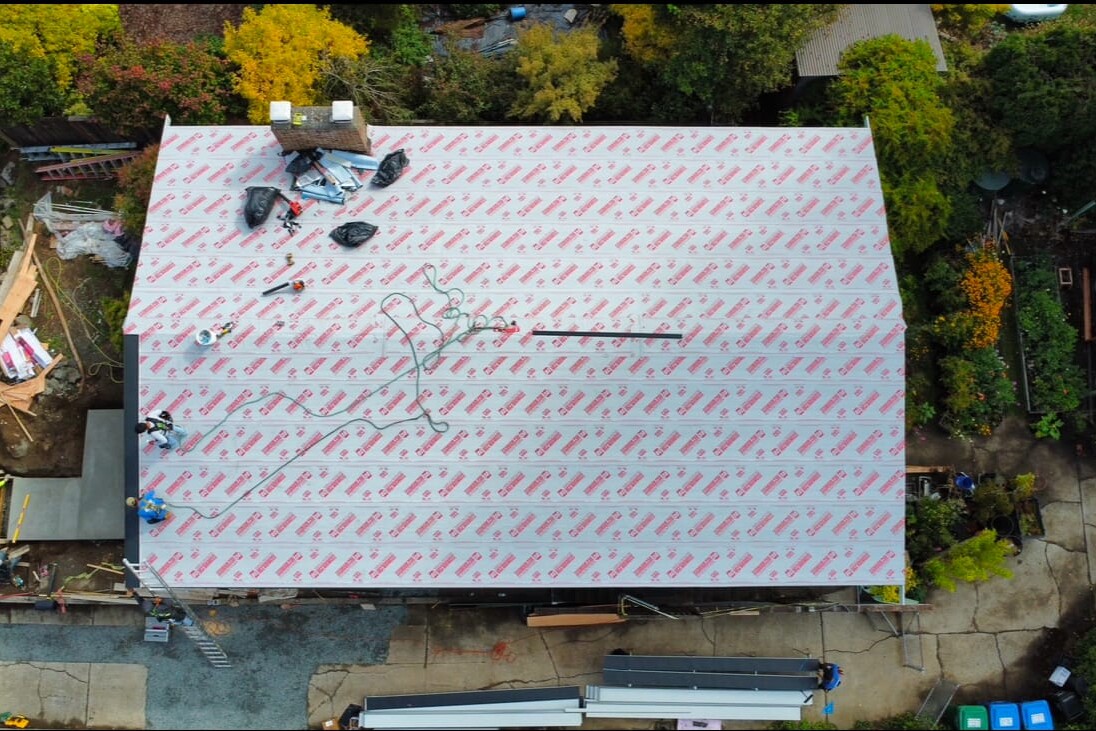 Aerial view of a houses rooftop under construction, covered in protective material with tools and equipment scattered. Workers are visible on the roof. Surrounding the house are trees with autumn-colored leaves and a few paths.