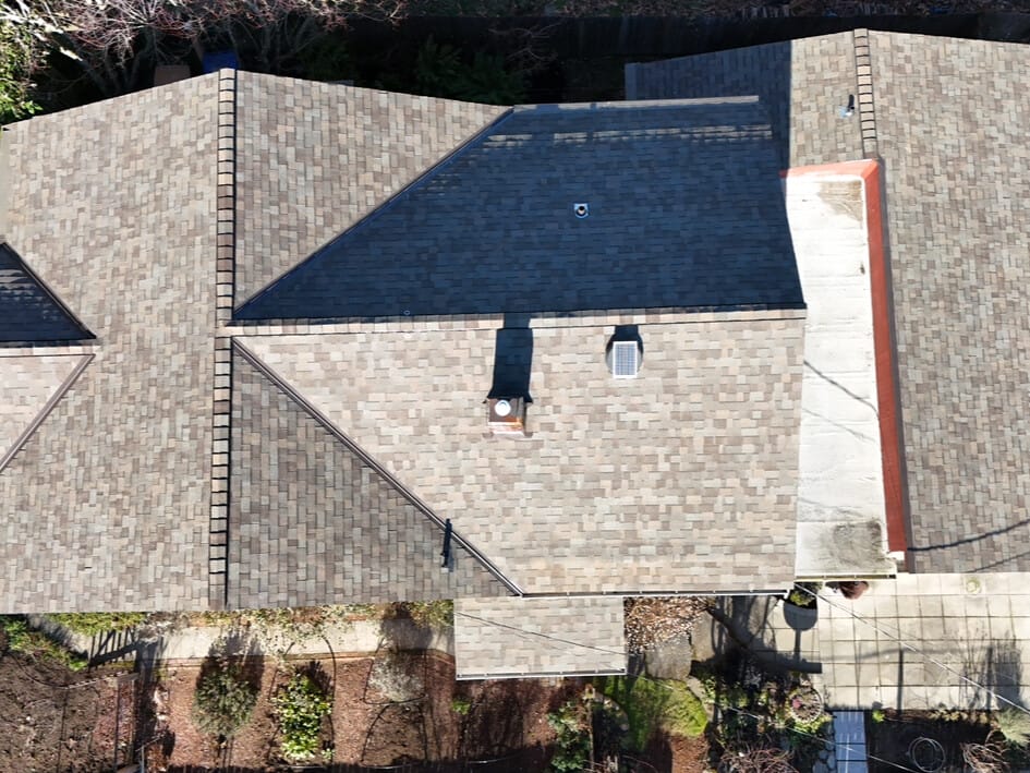Aerial view of a house with a brown shingle roof. The roof features multiple sections and a skylight. Surrounding the house are a lawn, paved pathways, and garden areas with sparse vegetation.