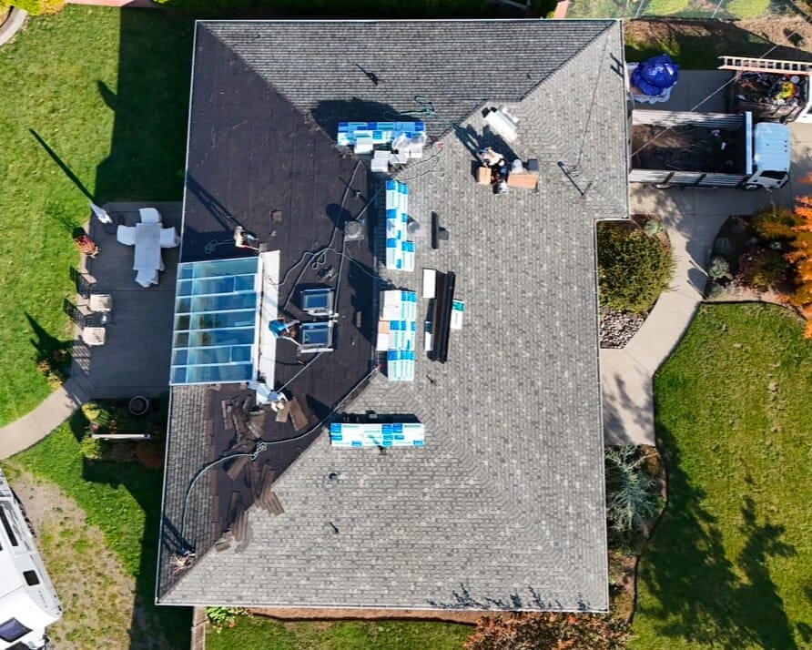 Aerial view of a house with solar panels and construction materials on the roof. The backyard features a patio with furniture and a small garden. Several vehicles, including a truck and an RV with solar panels, are parked nearby.