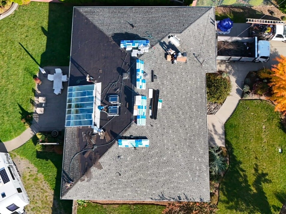 Aerial view of a house with solar panels and construction materials on the roof. The backyard features a patio with furniture and a small garden. Several vehicles, including a truck and an RV with solar panels, are parked nearby.