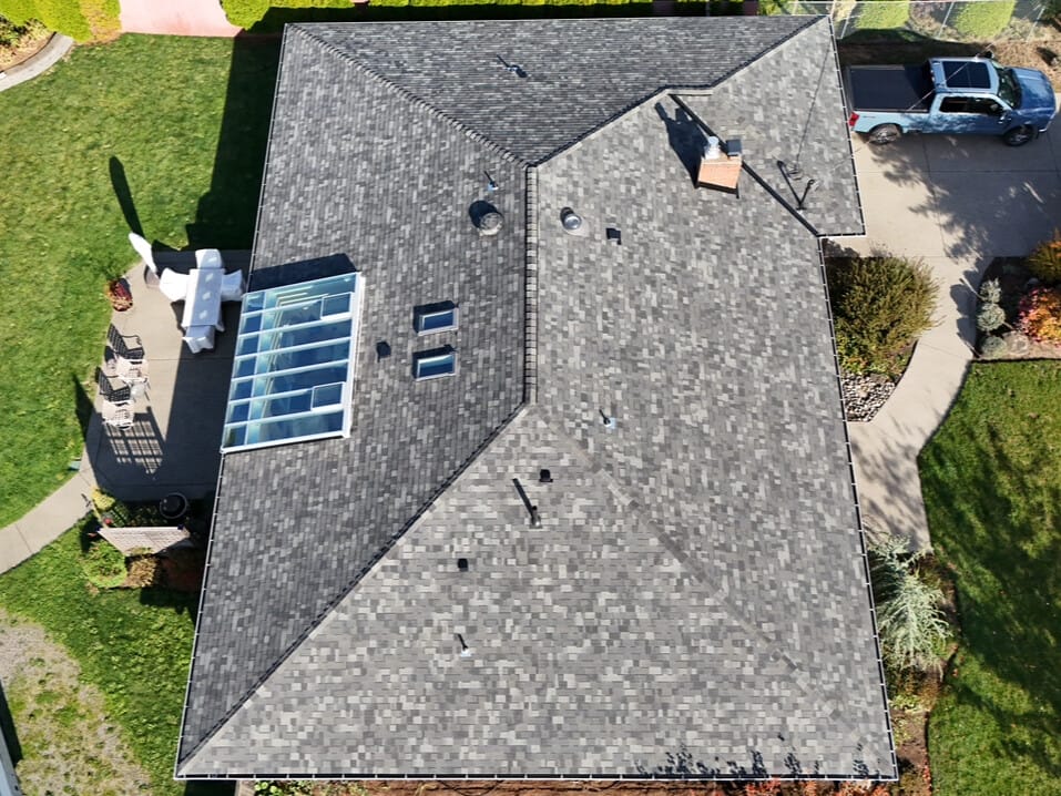 Aerial view of a house with a gray shingle roof featuring a skylight and chimney. The surrounding landscape includes a green lawn, driveway with a parked blue vehicle, and a patio area with outdoor furniture. Orange foliage is visible near the driveway.