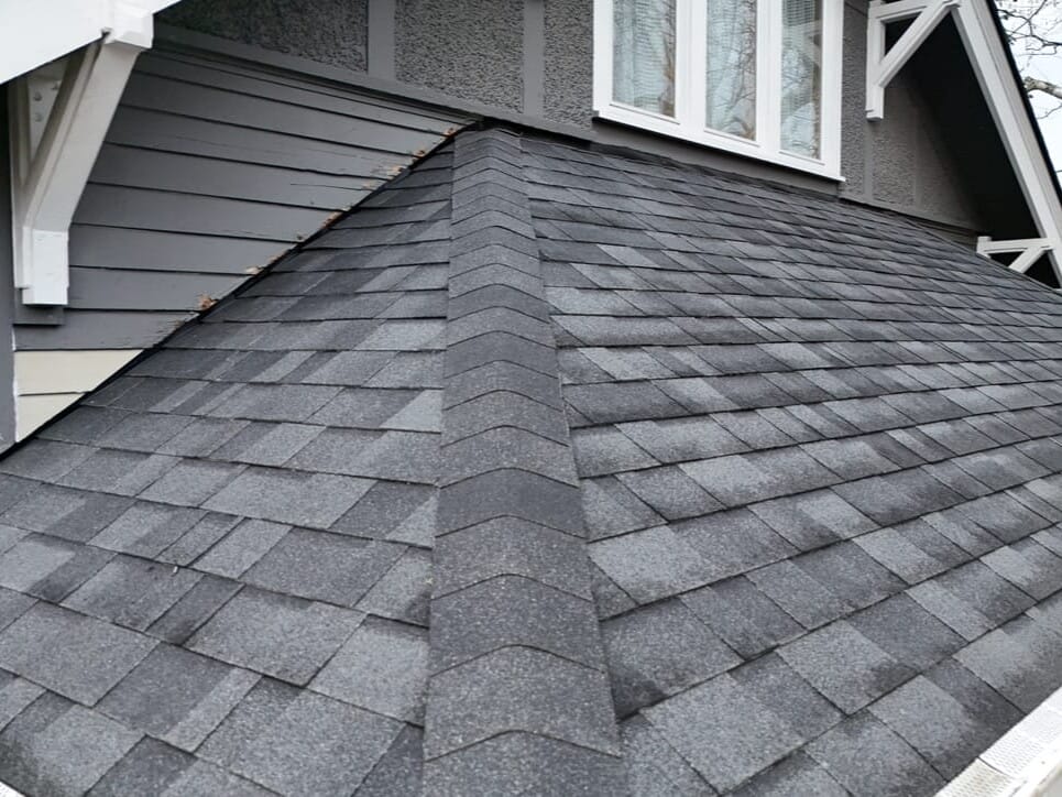 A close-up view of a house roof with dark gray asphalt shingles, featuring a ridge running down the center and white trim along the edges; the house exterior is painted gray with white window and eave accents.