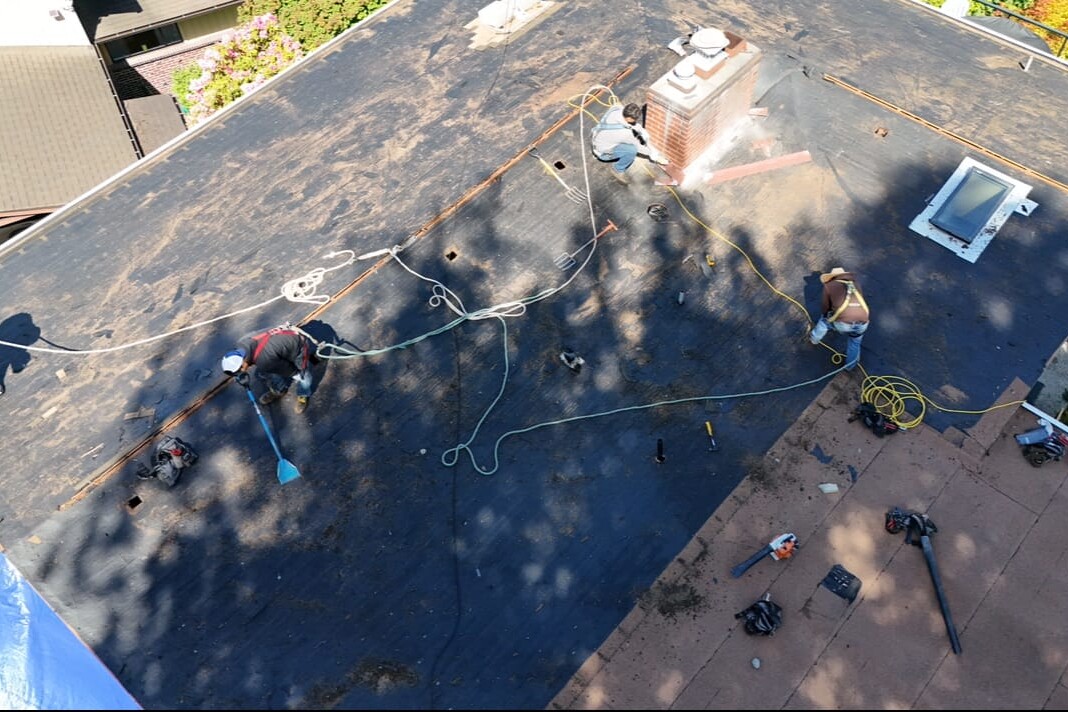 Aerial view of three workers on a flat roof installing or repairing roofing material, with tools, ropes, and equipment scattered around. There is a chimney and a skylight on the roof.