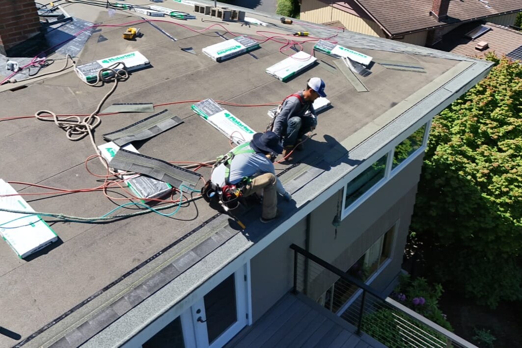Two workers wearing safety gear install shingles on a residential roof, surrounded by roofing materials, tools, and ropes, on a sunny day.