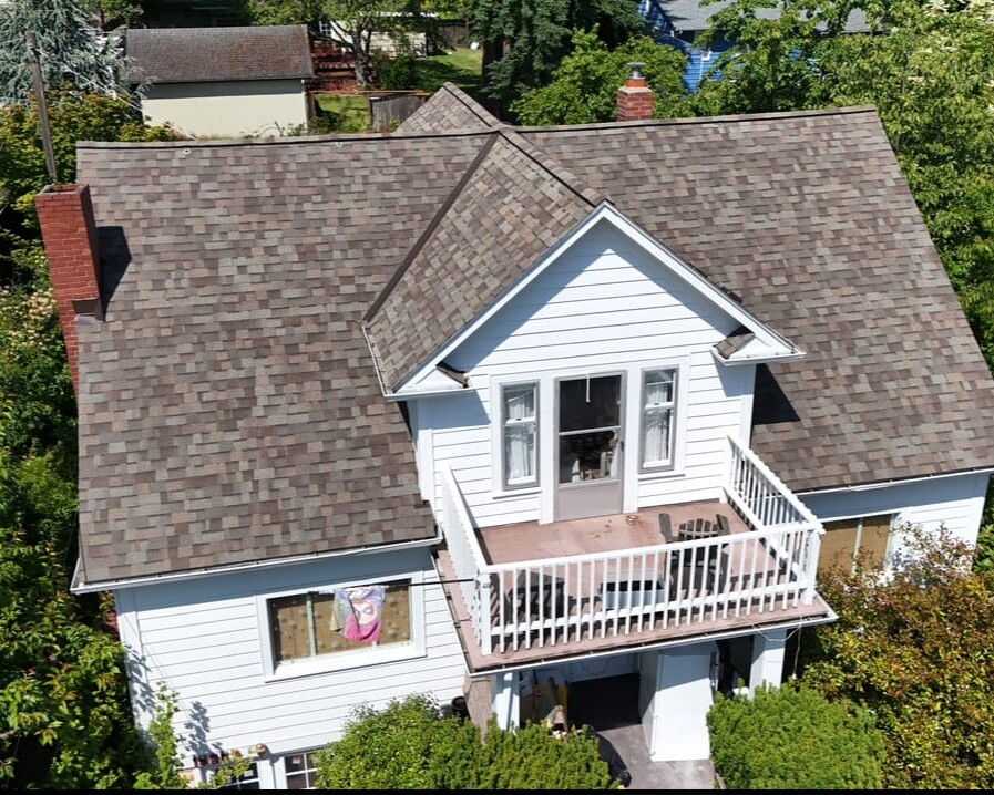 Aerial view of a two-story white house with a brown shingle roof, front balcony with a white railing, surrounded by trees and greenery. A window on the left has a large face image or poster on display.