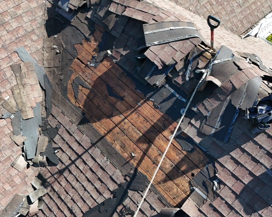 A damaged roof with missing shingles and exposed wooden decking, showing repair tools and materials nearby. A rope and a shovel are visible, indicating ongoing roof repair work.