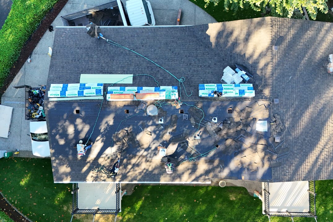 Aerial view of a house with workers installing new roofing. Shingles and materials are spread across the rooftop. Shadows of trees are cast on the roof, and a truck and garden are visible in the background.