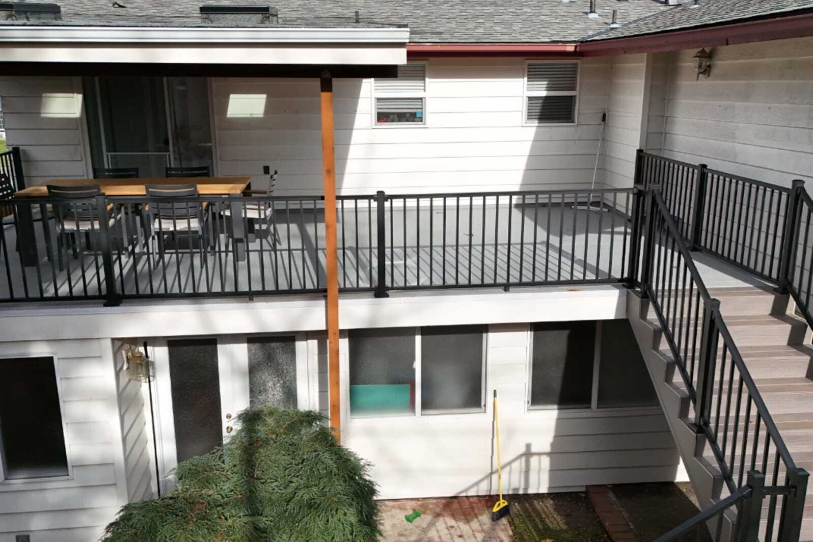 A two-story house with white siding and a black metal railing. The upper level features a patio with a table and chairs, and stairs lead from there to the lower level. A broom leans against the wall in the lower area.
