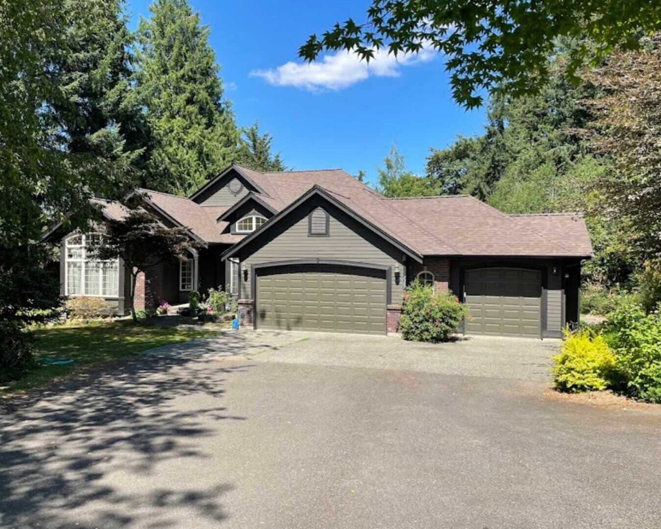 A single-story house with dark gray siding and a brown roof, featuring three garage doors. Its surrounded by lush greenery and tall trees under a clear blue sky. A paved driveway leads up to the house.