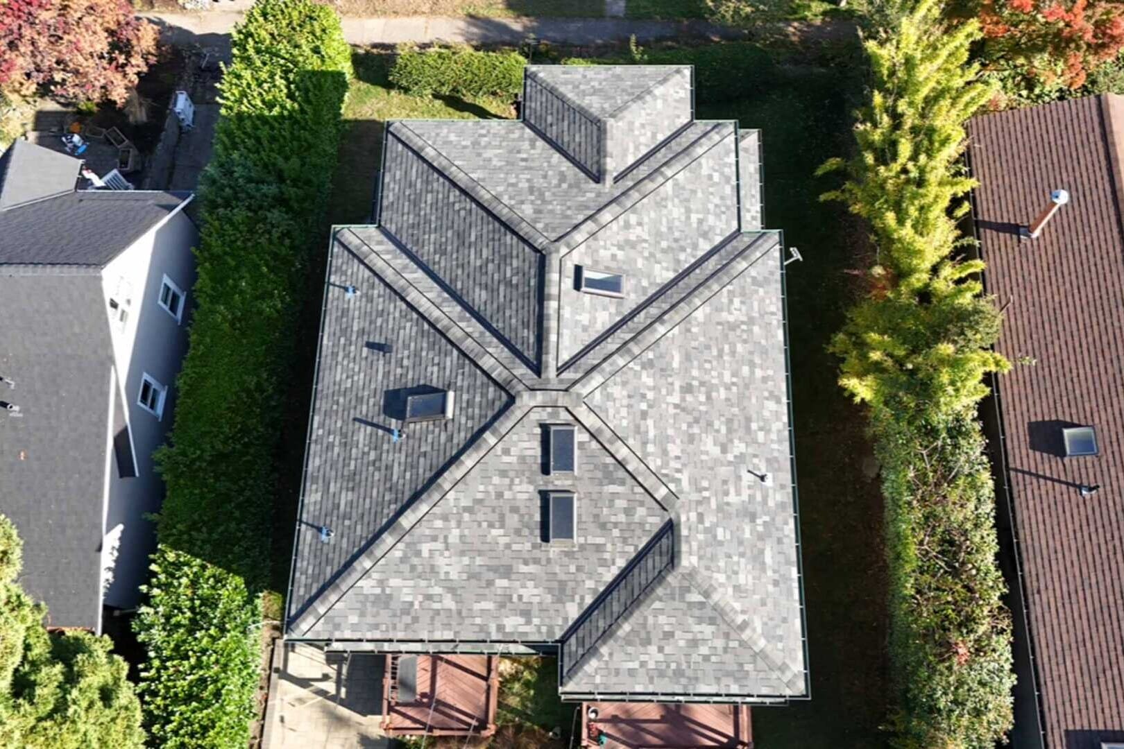Aerial view of a large house with a complex, geometric gray shingled roof. The house is flanked by trees and is situated between two neighboring houses. The yard is partially visible with greenery surrounding the property.