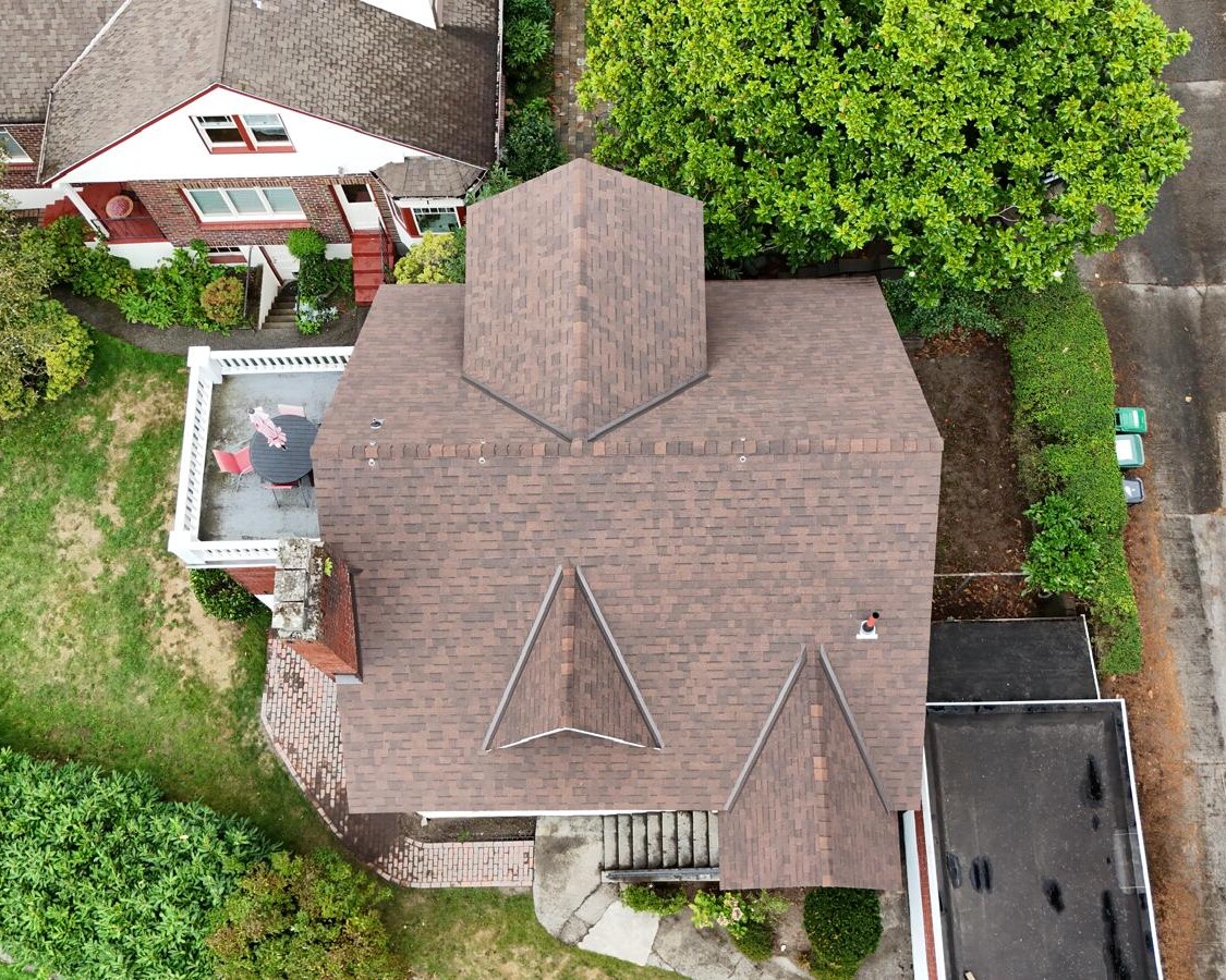 Aerial view of a brown-roofed house with distinctive triangular designs. A person stands on a small rooftop patio adorned with an American flag. Surrounding the house are trees, a garden, and adjacent driveways. Another house with a similar design is nearby.