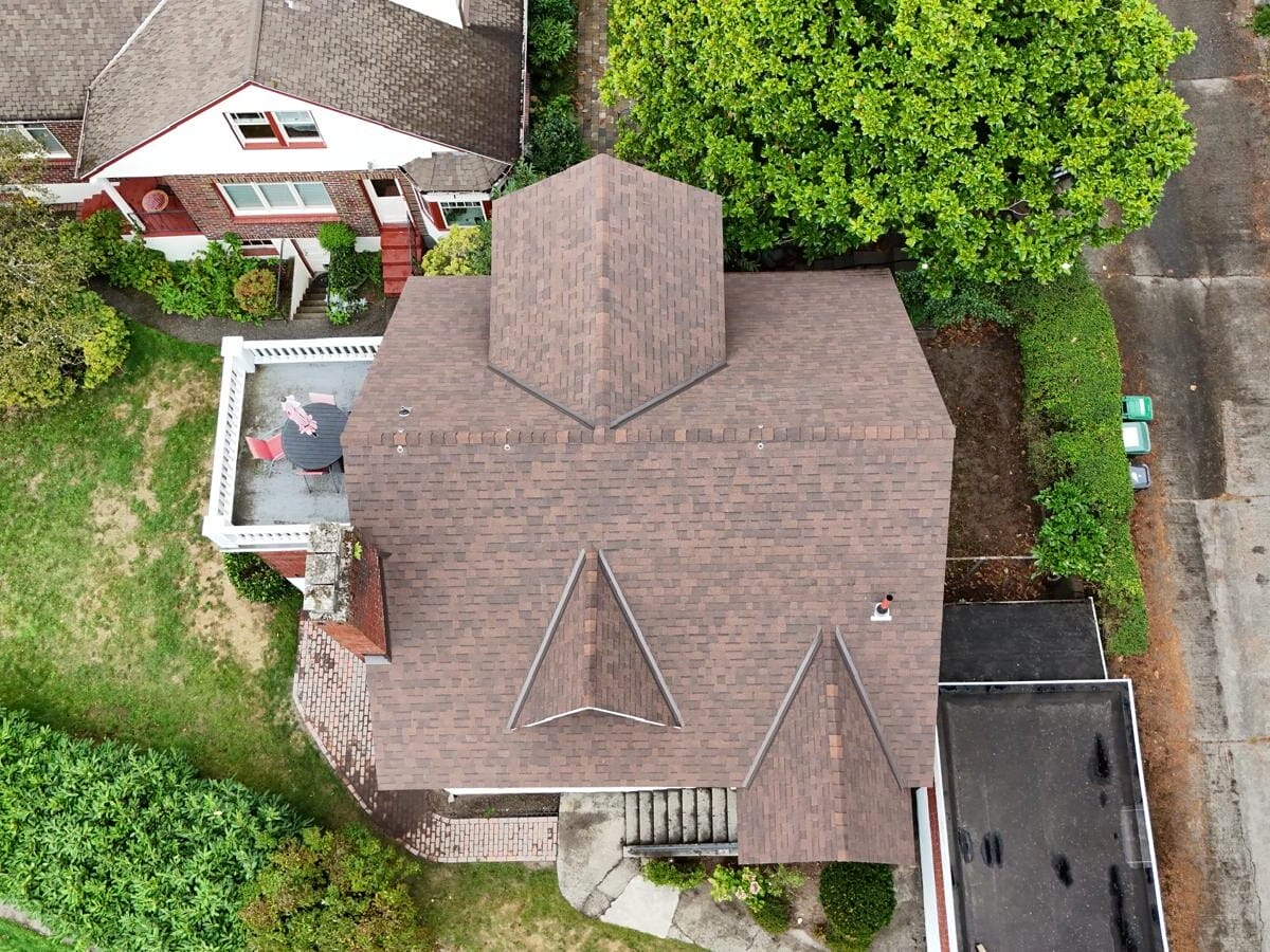 Aerial view of a brown-roofed house with distinctive triangular designs. A person stands on a small rooftop patio adorned with an American flag. Surrounding the house are trees, a garden, and adjacent driveways. Another house with a similar design is nearby.