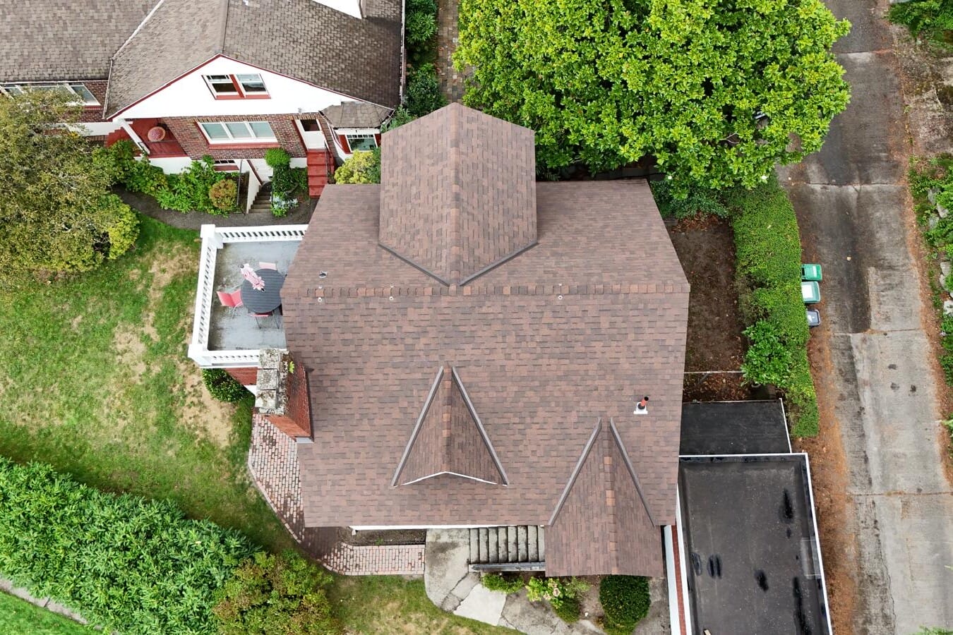 Aerial view of a brown-roofed house with distinctive triangular designs. A person stands on a small rooftop patio adorned with an American flag. Surrounding the house are trees, a garden, and adjacent driveways. Another house with a similar design is nearby.