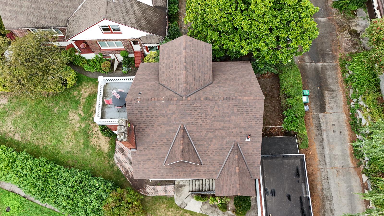 Aerial view of a brown-roofed house with distinctive triangular designs. A person stands on a small rooftop patio adorned with an American flag. Surrounding the house are trees, a garden, and adjacent driveways. Another house with a similar design is nearby.