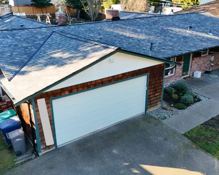 Aerial view of a single-story suburban house with a two-car garage, shingle roof, brick and wood siding, concrete driveway, and a walkway leading to the front entrance.