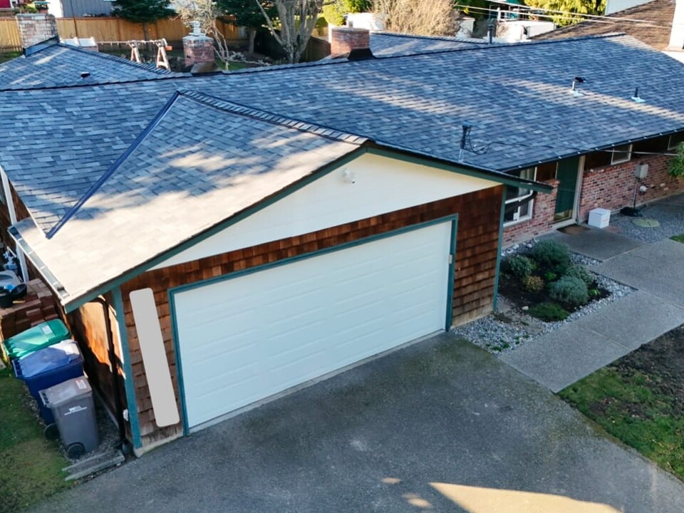 Aerial view of a single-story suburban house with a two-car garage, shingle roof, brick and wood siding, concrete driveway, and a walkway leading to the front entrance.