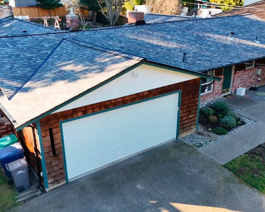 A single-story house with a shingle roof, a white garage door, brown wooden siding, and a concrete driveway. There are trash bins on the side and a small landscaped area with shrubs near the entrance.