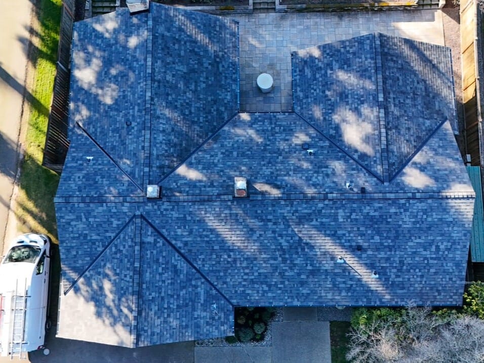 Aerial view of a building with a dark gray, multi-pitched roof. A white vehicle with a ladder is parked on the left side. The surrounding area includes roads and trees with long shadows cast on the ground.
