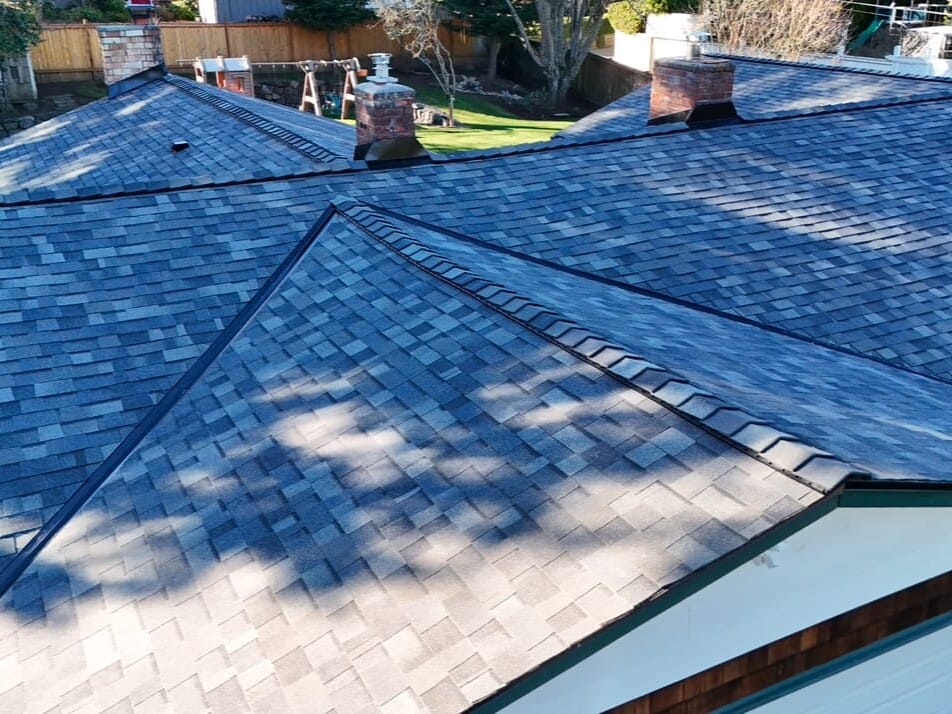 Aerial view of a house with a newly installed, dark gray shingle roof. The roof features multiple triangular sections and ventilation outlets. Surrounding trees cast shadows on the roof, and a backyard with grass is partially visible.