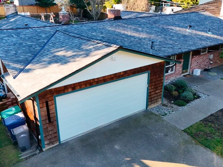 Aerial view of a single-story house with a dark shingle roof. The house has a white garage door and brick walls. A driveway leads to the garage, and there is a small, landscaped garden area next to a sidewalk.