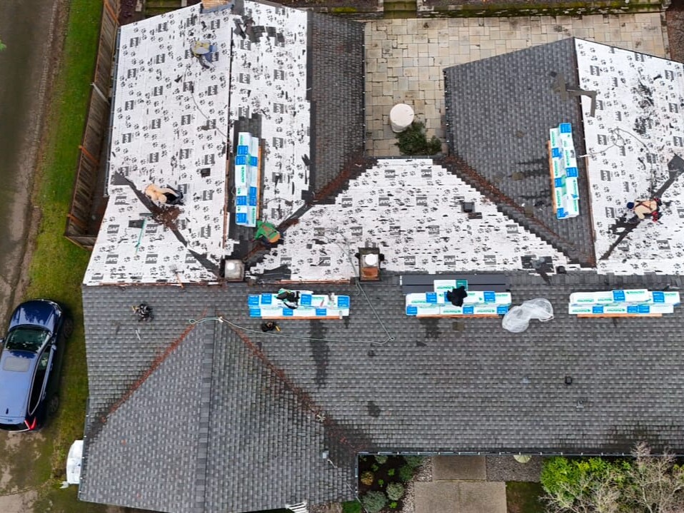 Aerial view of workers installing new roofing material on a house. Some shingles are already in place, while bundles of shingles and tools are spread across the roof. A blue car is parked in the driveway beside the house.