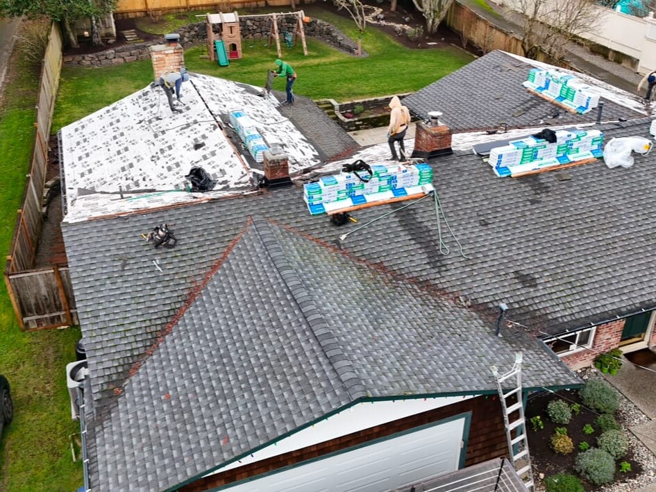 Aerial view of workers installing new shingles on a house roof, with roofing materials stacked in bundles. Some sections of the roof are bare or covered in underlayment, while others have finished shingles.