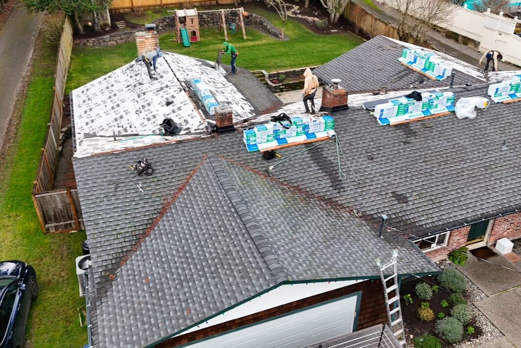Aerial view of workers on a house roof installing new shingles. The roof is partially covered, with stacks of materials visible. Surrounding the house are a lawn, a driveway with a car, and a tree-lined fence.