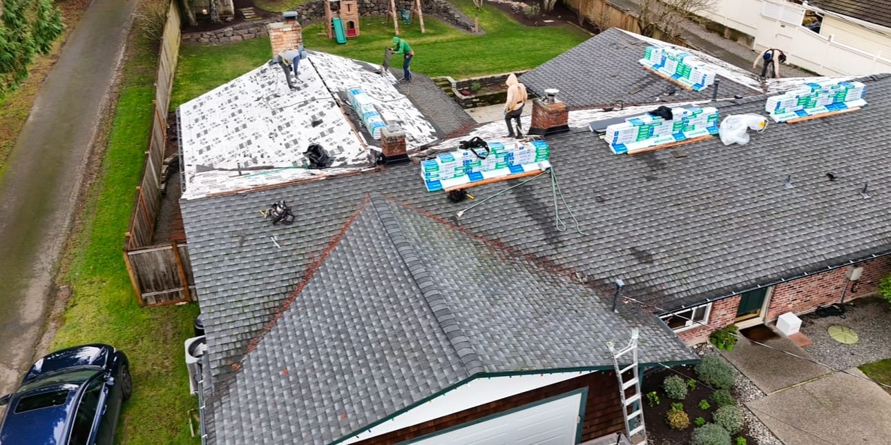 Aerial view of workers on a house roof installing new shingles. The roof is partially covered, with stacks of materials visible. Surrounding the house are a lawn, a driveway with a car, and a tree-lined fence.