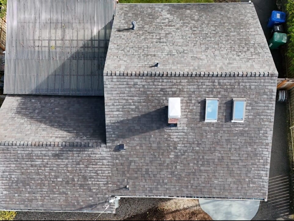 Aerial view of a houses roof with dark shingles. The roof has multiple sections and includes three skylights and a chimney. There are green and blue trash bins along a pathway beside the house, lined with trees and a fence.