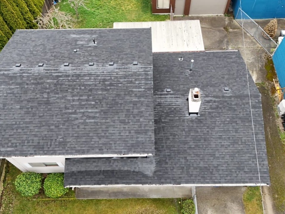 Aerial view of a house with a dark gray shingle roof. The house is surrounded by green grass, with a few bushes and trees nearby. Adjacent buildings include a blue structure and a garage. Theres a chimney on the roof.