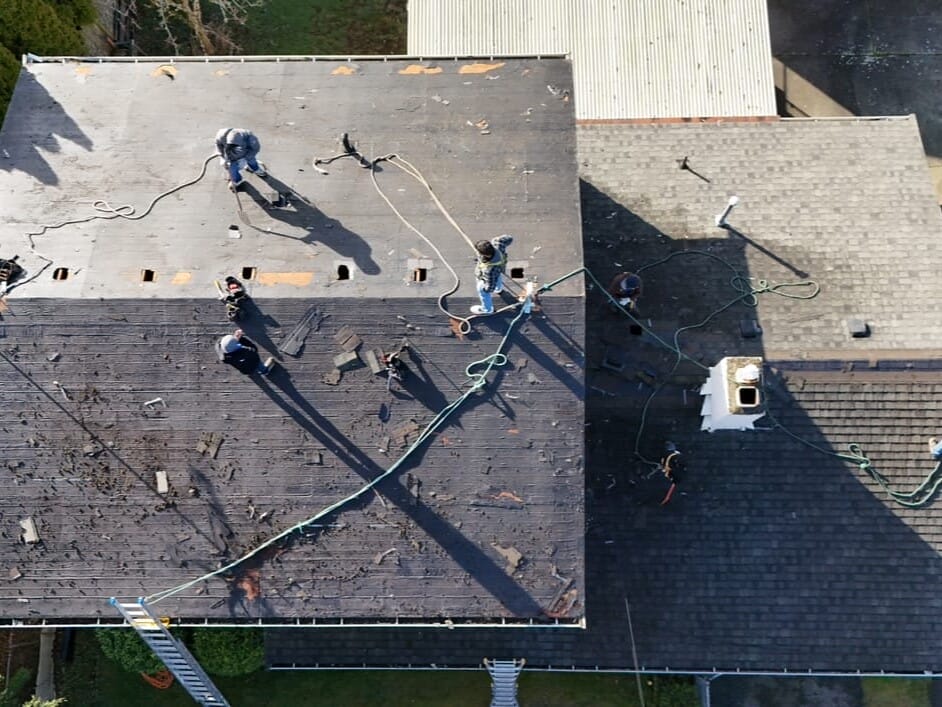 Aerial view of several workers on a roof removing shingles. The roof is partially stripped, revealing the underlying structure. Ladders and ropes are visible, and a chimney is on the right side. Surrounding areas include trees and buildings.