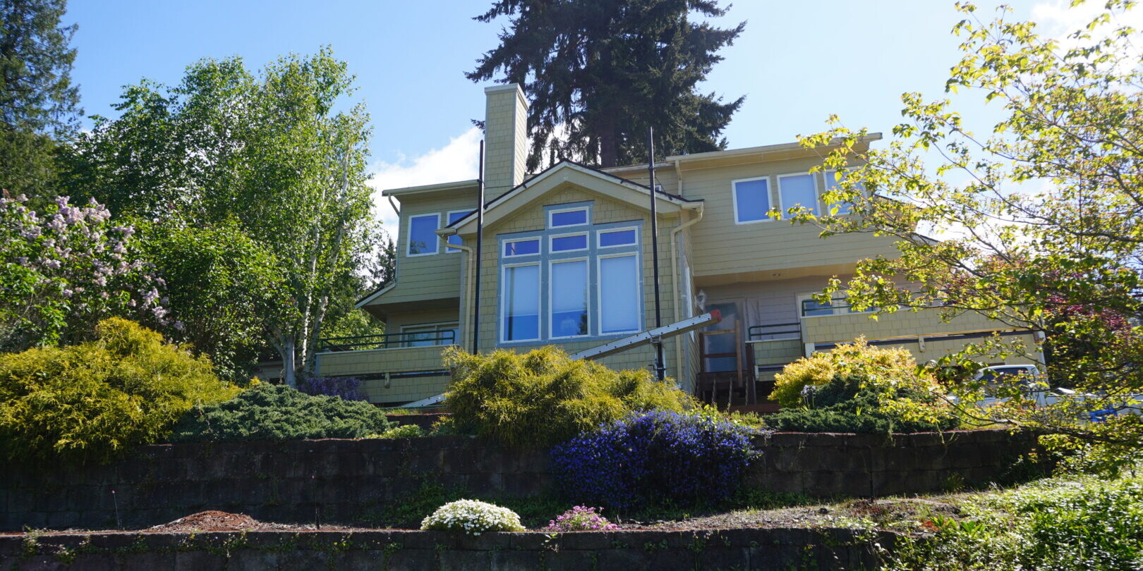 A yellow two-story house with a large tree behind it. The house has multiple windows and a chimney, surrounded by a garden with shrubs and blooming flowers. A stone retaining wall with greenery tiers up to the house.