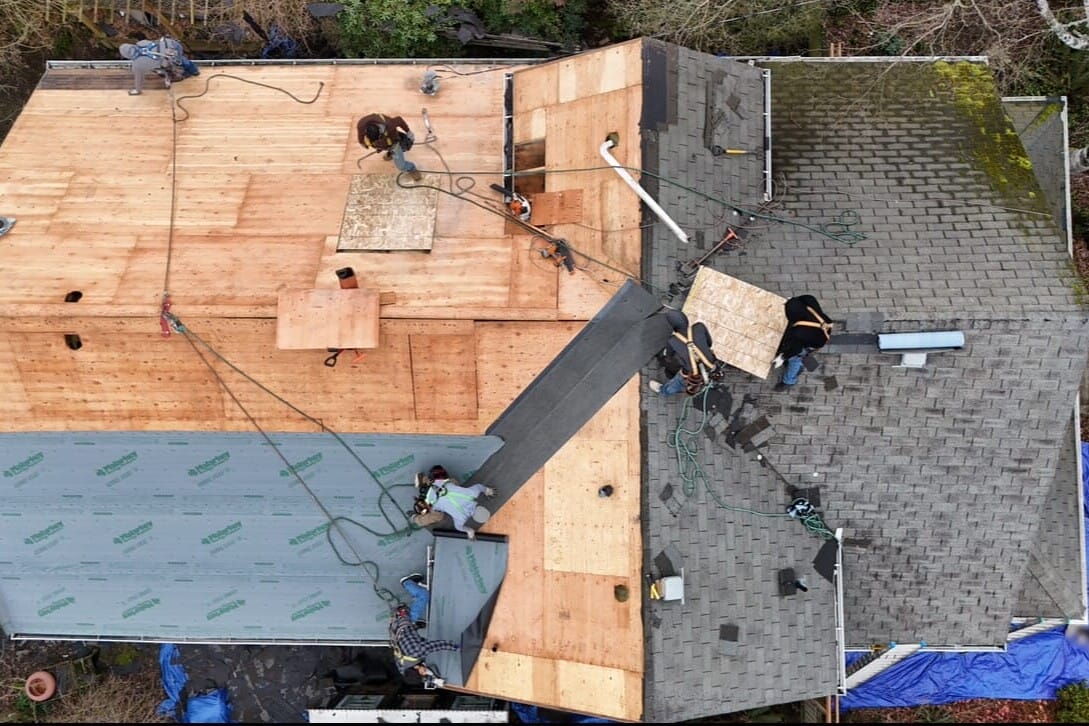 Aerial view of workers replacing a house roof; half of the roof shows new plywood, while the other half has old gray shingles. Ropes and equipment are visible, with workers actively installing materials.